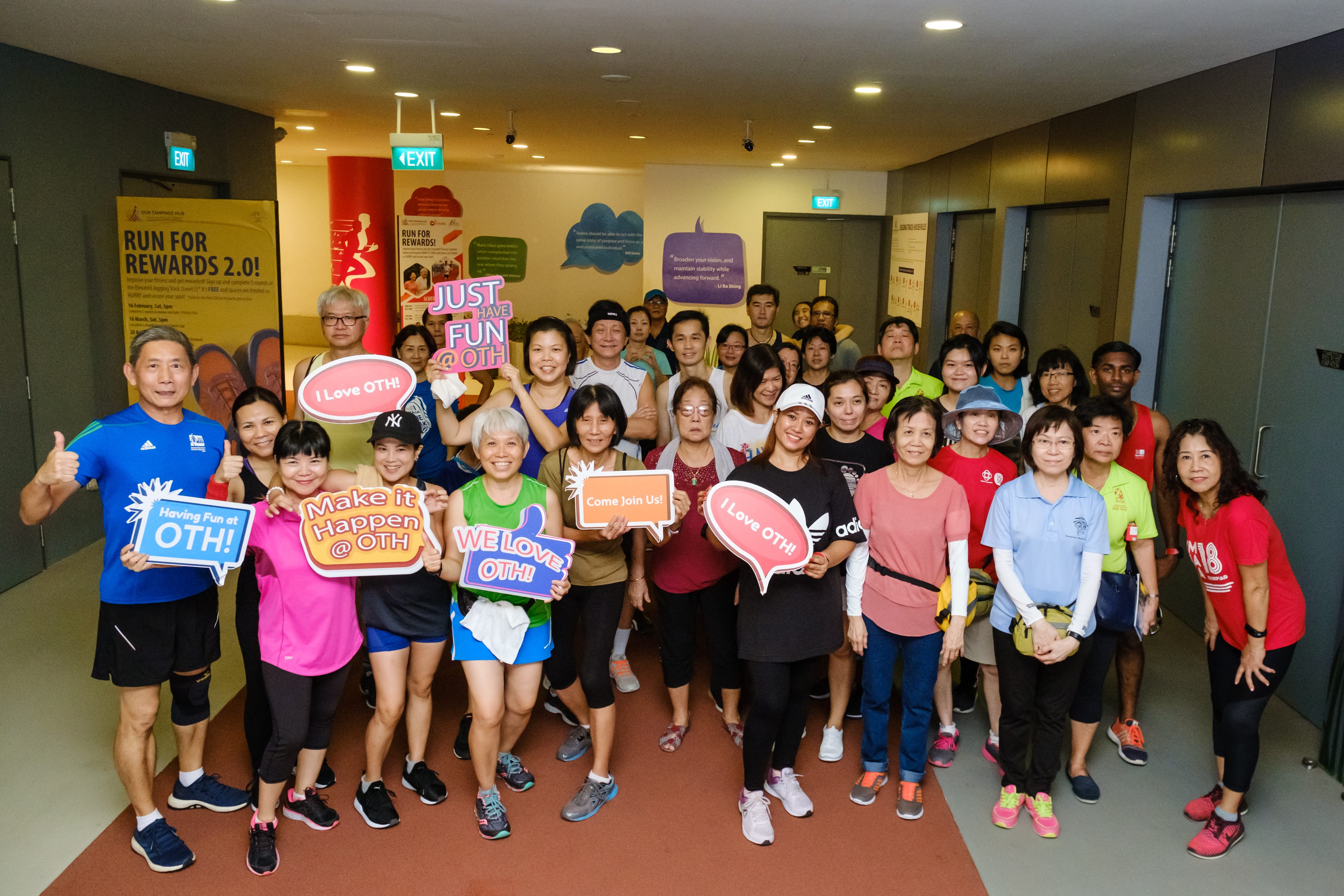 A large group of people stand indoors holding colorful signs, including "Make it Happen @OTH" and "We Love OTH!".